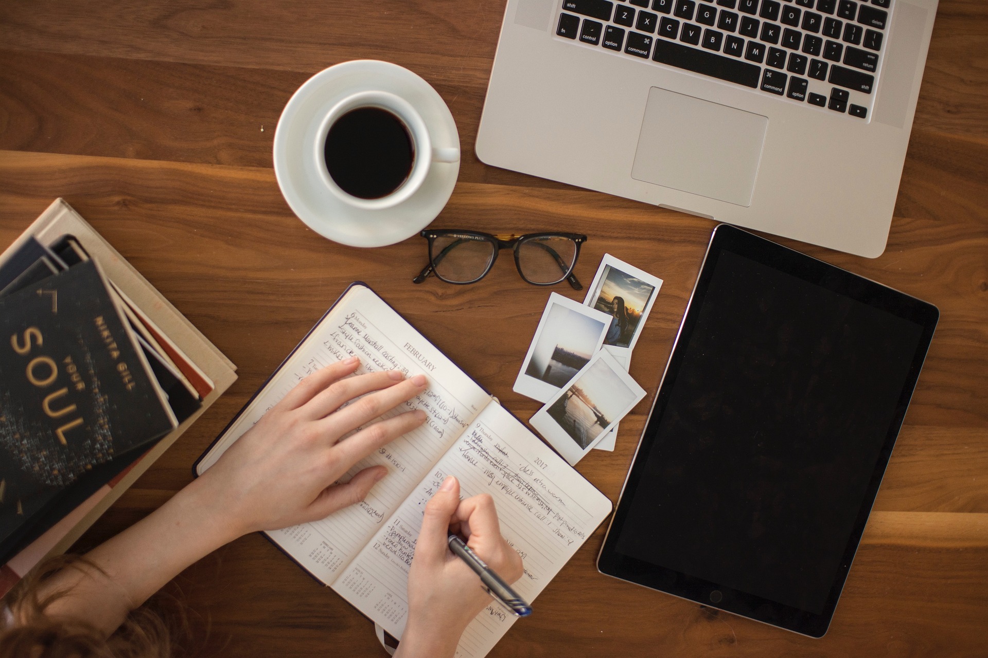 Eine Person schreibt mit einem Stift Ideen in ein offenes Notizbuch, umgeben von einem Laptop, einer Tasse Kaffee, einer Brille und Büchern, die Inspiration bieten.